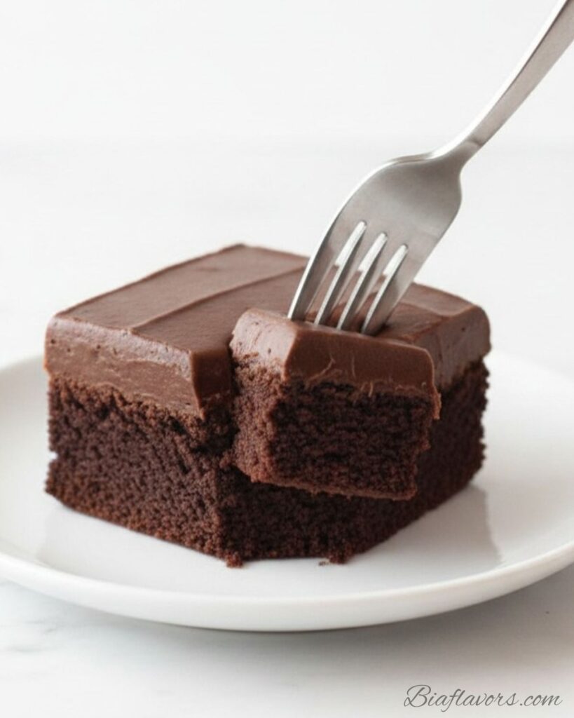 _Silver fork cutting into frosted fudge brownie on white plate lifting bite showing dense dark fudgy interior with thick chocolate frosting on top