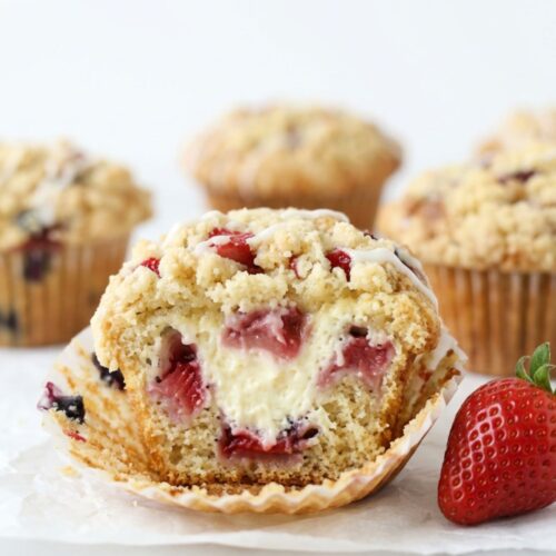 Dramatic close-up of strawberry cheesecake streusel muffin with wrapper peeled showing creamy cheesecake filling strawberry chunks golden streusel and white glaze drizzle