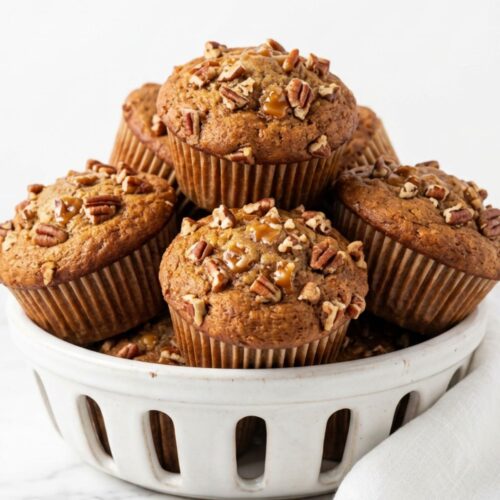 Pile of caramel banana nut muffins with chopped pecans on top stacked in white ceramic basket on white marble counter clean white background