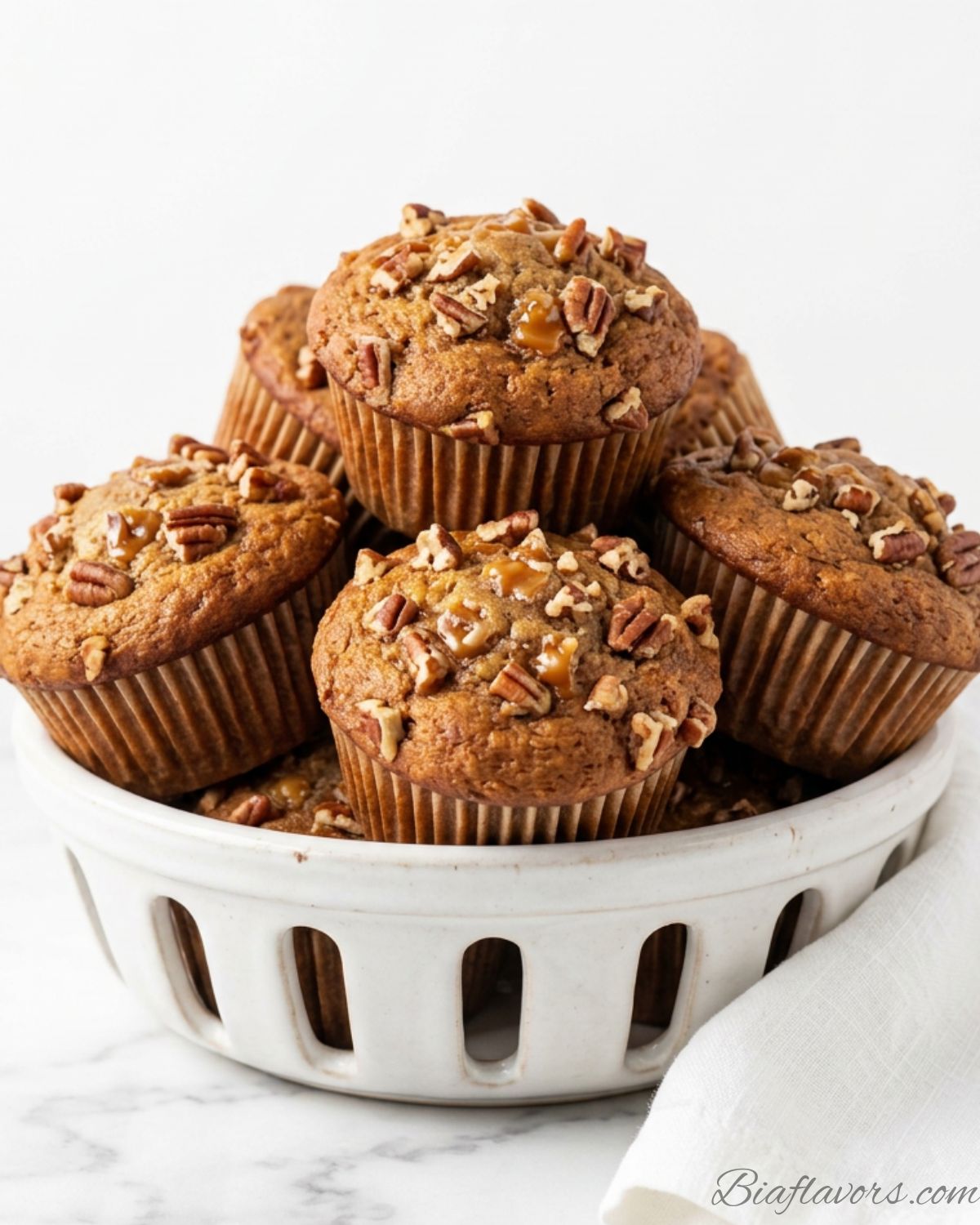 Pile of caramel banana nut muffins with chopped pecans on top stacked in white ceramic basket on white marble counter clean white background