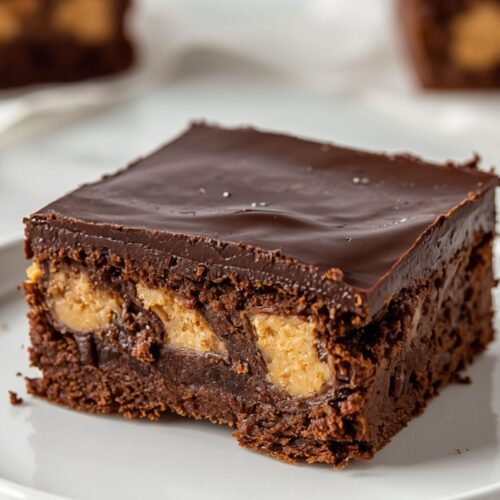 Single peanut butter stuffed brownie square on white plate showing glossy chocolate ganache top and visible peanut butter chunks inside on white marble