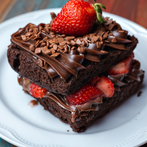 Dramatic close-up of fudgy strawberry brownie on white plate showing dark chocolate layers ganache filling sliced strawberries mini chips and whole strawberry on top