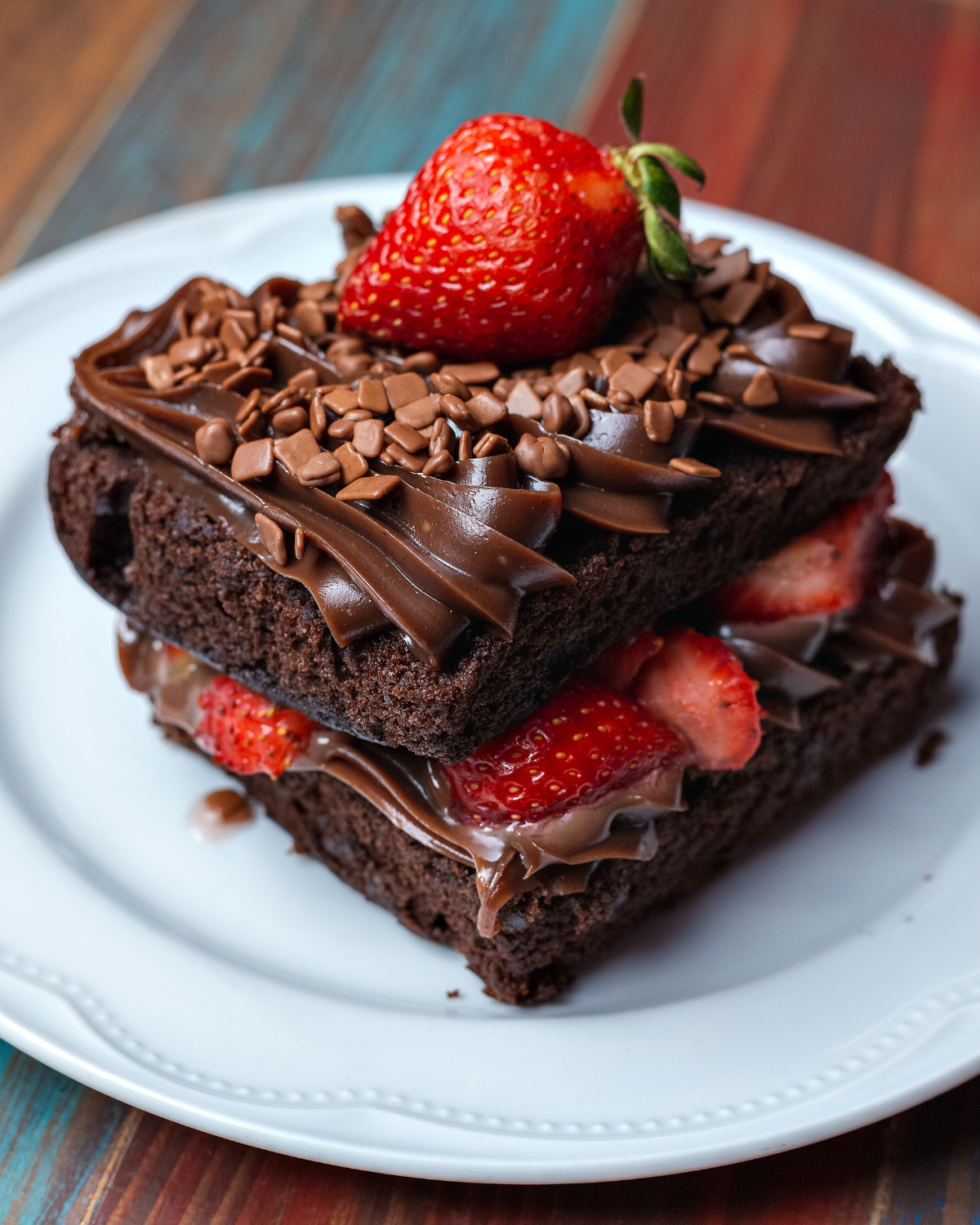 Dramatic close-up of fudgy strawberry brownie on white plate showing dark chocolate layers ganache filling sliced strawberries mini chips and whole strawberry on top